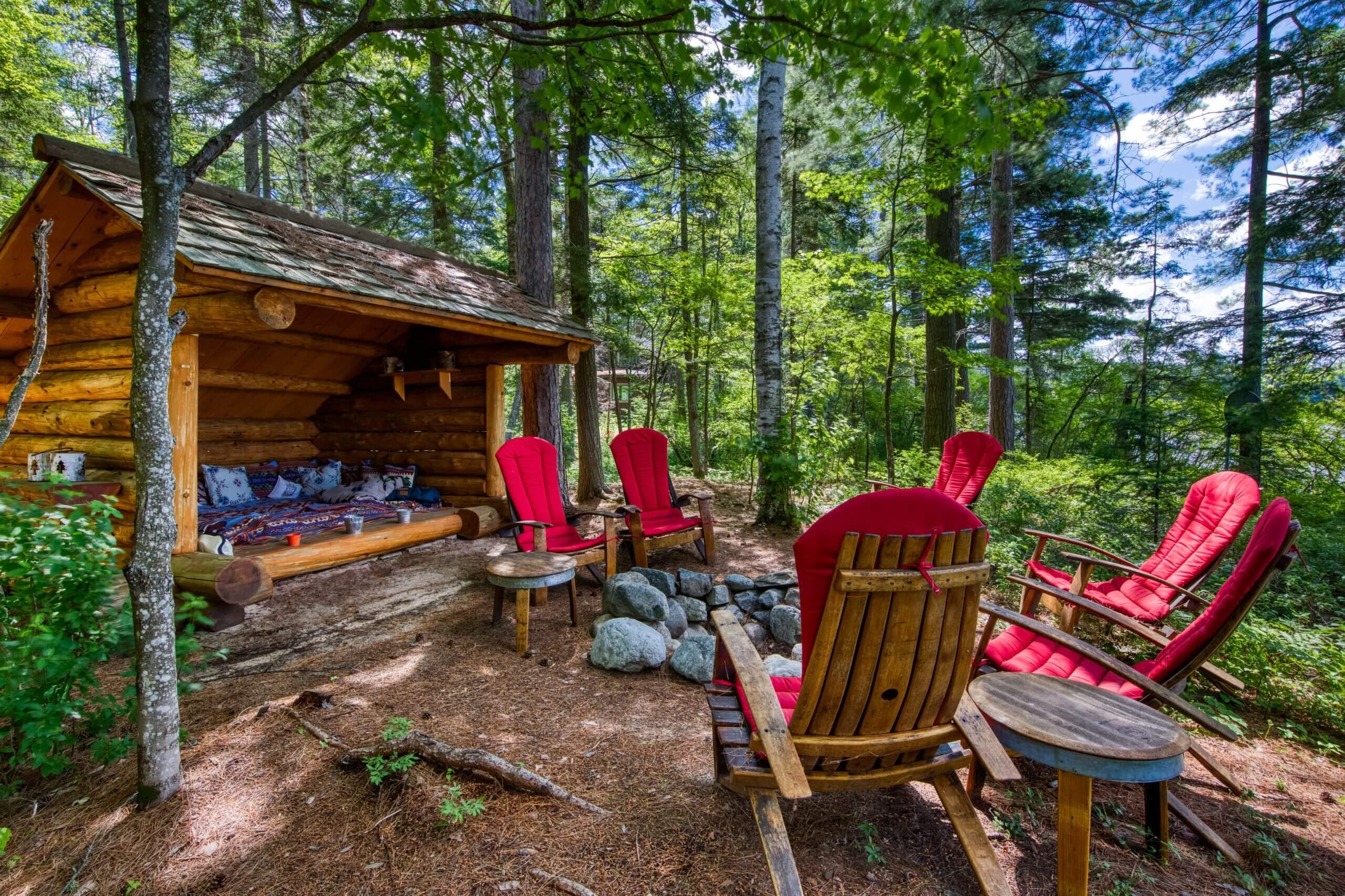 A furnished lean to on a vacation rental property in the Adirondacks with a stone fire ring and red Adirondack chairs surrounding it