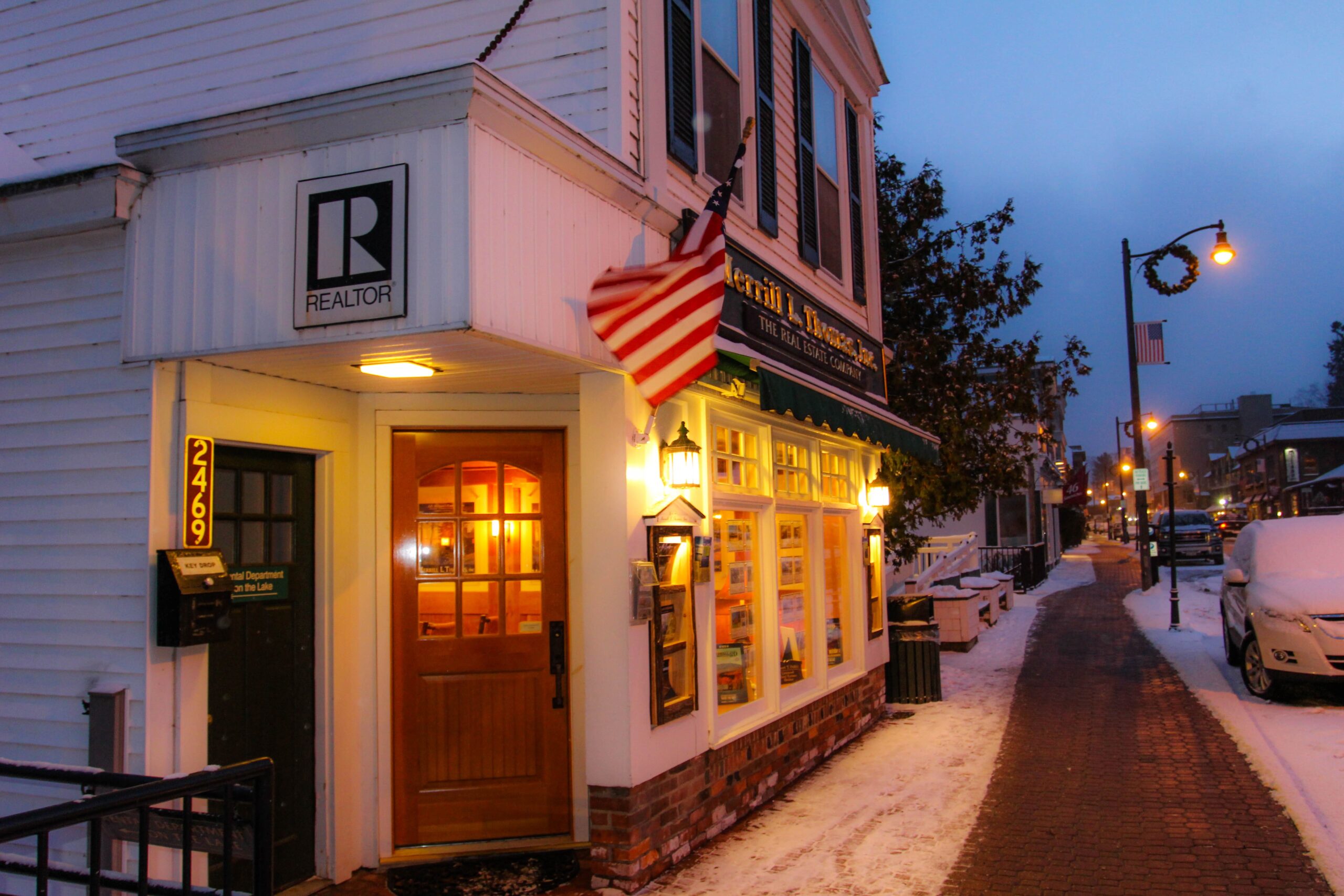 Nighttime shot of Merrill Thomas's Lake Placid office with lights on inside and a flag waving in front.